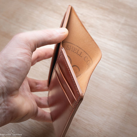 Hand holding a Shell Cordovan brown leather wallet with 'Explorer Leather' branding on a wooden surface