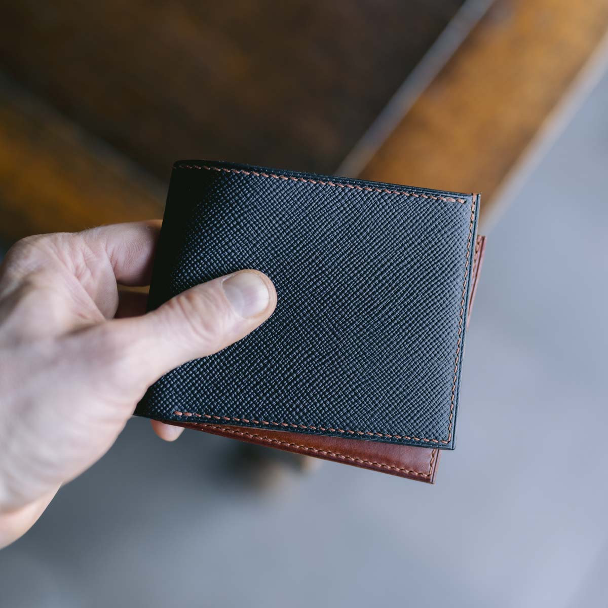 Hand holding a black and brown leather wallet with brown stitching on a blurred background