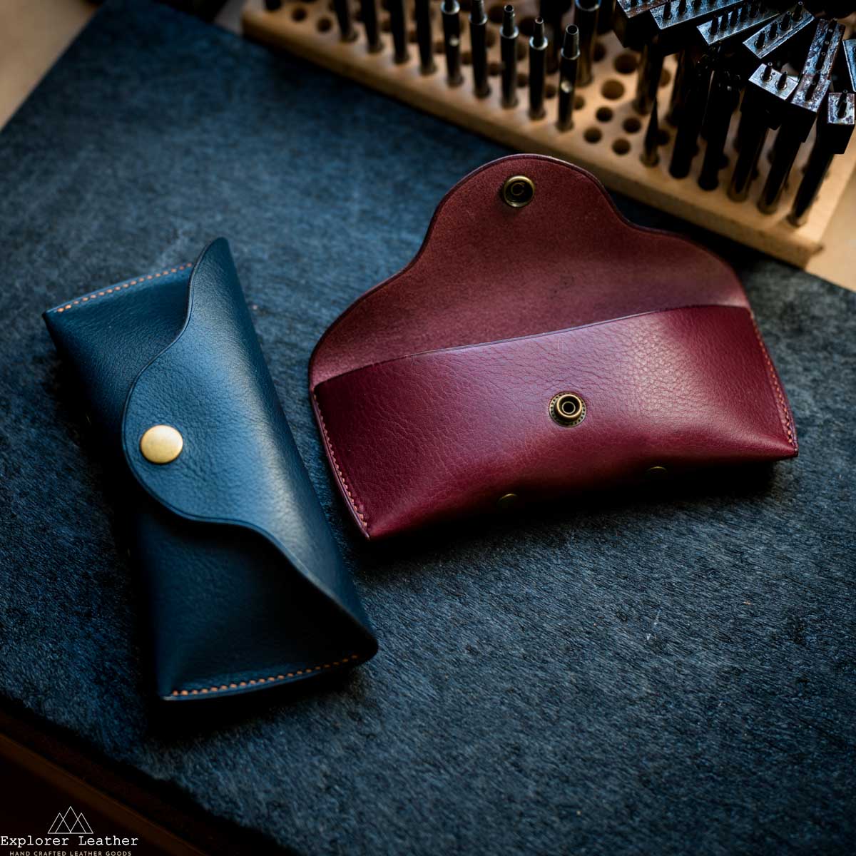 Two leather sunglasses cases, one blue and one burgundy, on a textured surface with 'Explorer Leather' branding.