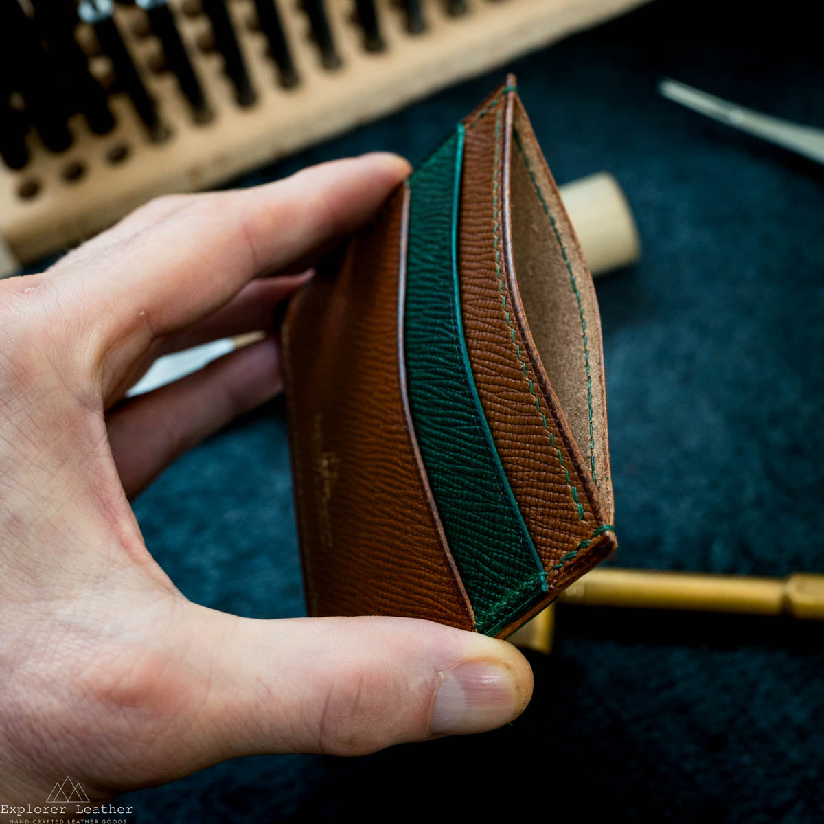 Hand holding a brown leather wallet with green stitching on a dark textured background.