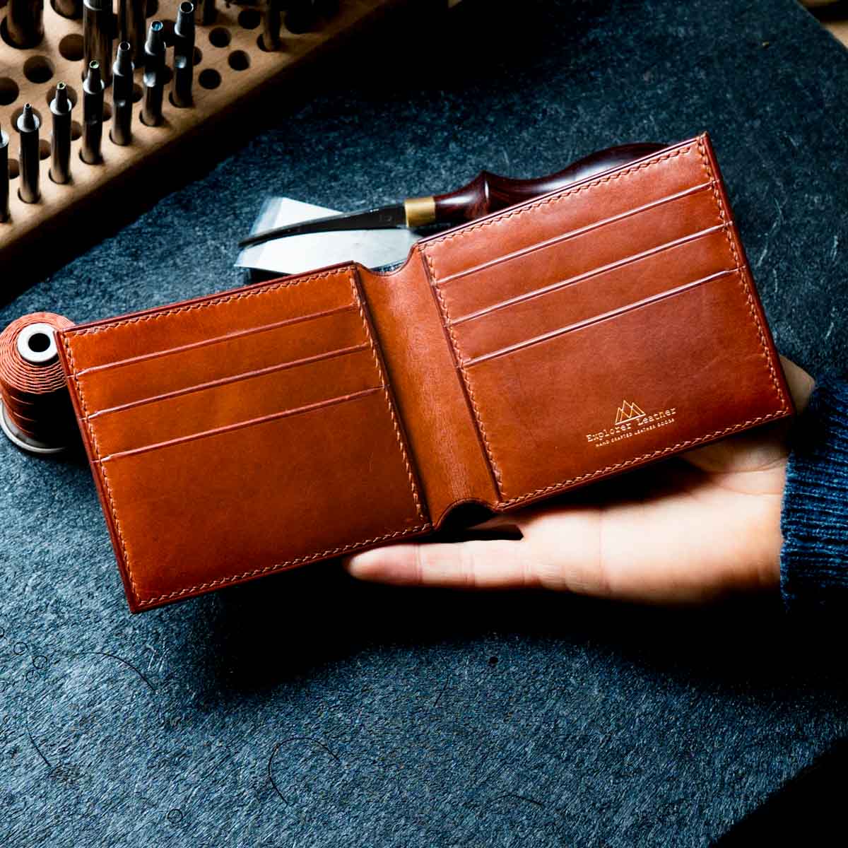 Brown leather wallet held by a hand on a dark textured surface with tools in the background.
