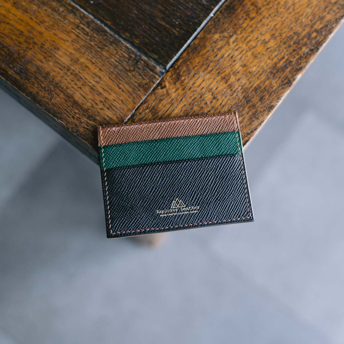 Black, green and brown leather cardholder with a visible explorer leather brand logo. On a wooden surface