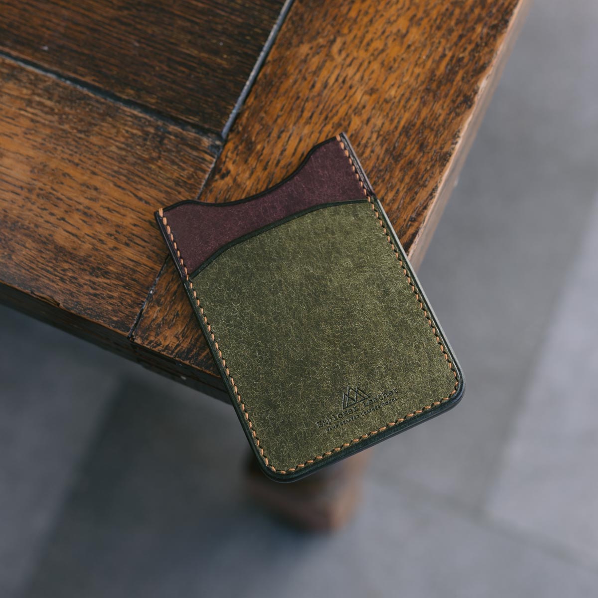 Hand holding a Green and Brown leather cardholder on a wooden surface.