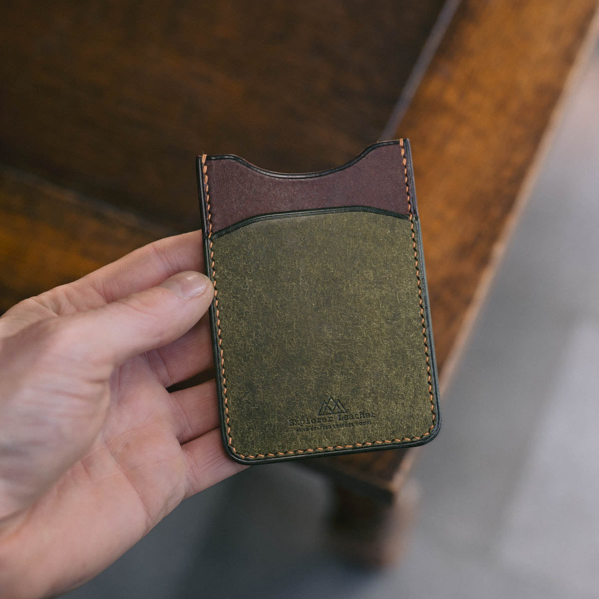 Hand holding a Green and Brown leather cardholder on a wooden surface.